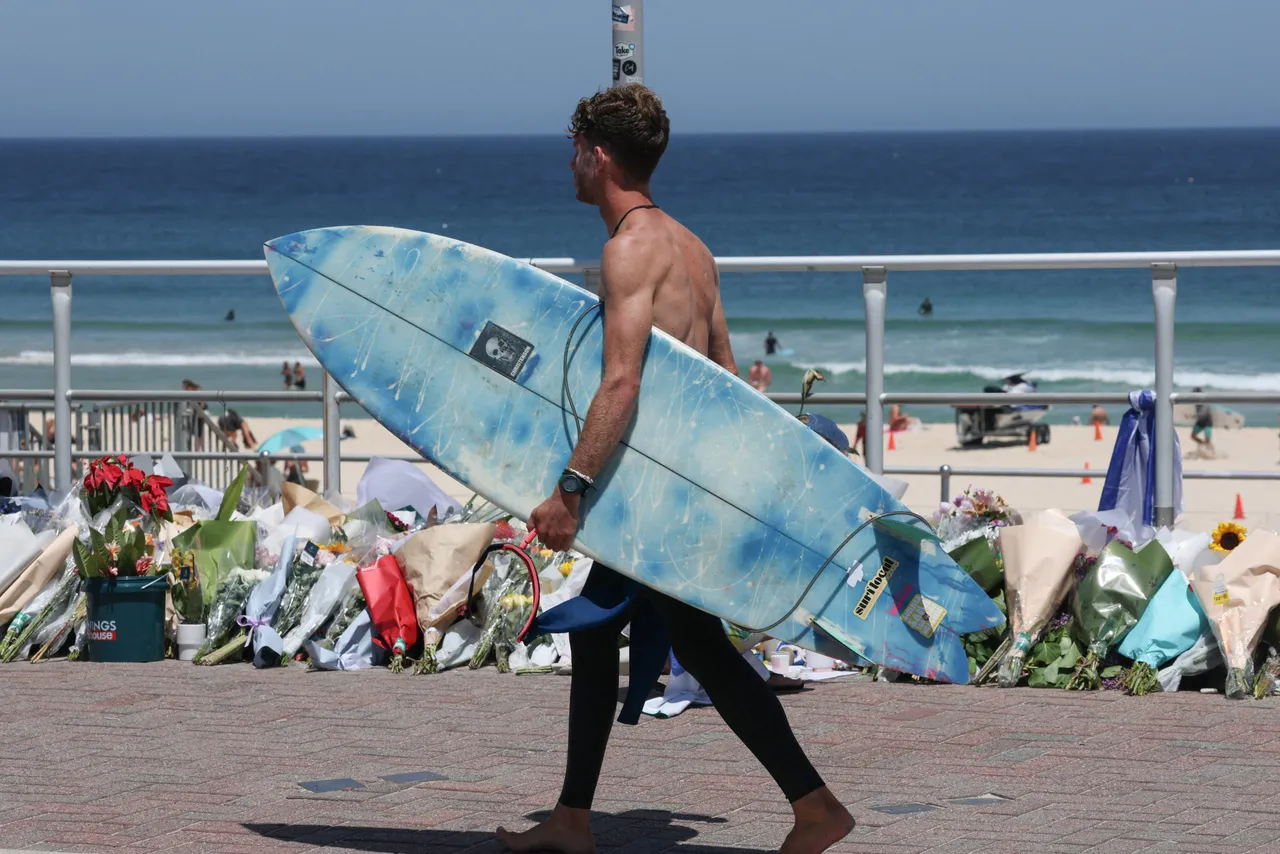A surfer walks past floral tributes left at the promenade of Bondi Beach in Sydney on December 18, 2025, to honour victims of the shooting that took place there on December 14. The attack at Bondi Beach on December 14 was one of the deadliest in Australian history. (Photo by DAVID GRAY / AFP via Getty Images)